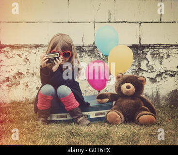 A photo of a vintage child taking a picture with old camera against a brick wall, balloons and a teddy bear for a creative idea