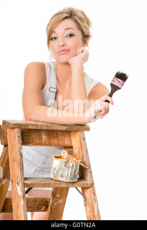 Cute blonde girl, in overalls, holding paint brush and resting on old ladder Stock Photo