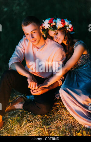 Beautiful girl in dress with wreath putting her head on the shoulder of nice guy sitting down outdoors at night Stock Photo