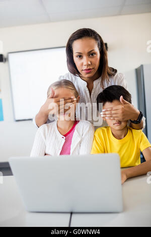Teacher covering pupils eyes in front of computer and looking at camera Stock Photo