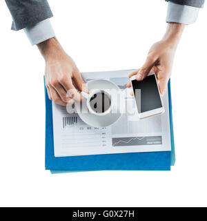 Businessman having a coffee break and using a smart phone, hands close up, top view Stock Photo