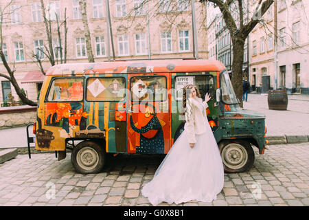 Stylish bride in white dress and fur cape smiling having fun near colorful retro car Stock Photo