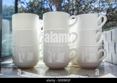 White cups for coffee and tea piled on vintage table close to window open to garden area Stock Photo