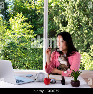 Mature woman, wearing pink bathrobe, holding her family cat while working from home in front of large window Stock Photo