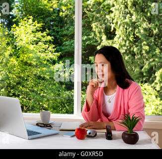 Mature woman, wearing pink bathrobe, working from home in front of large window with bright daylight and trees in background. Stock Photo