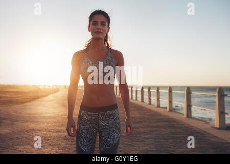 Portrait of confident young sportswoman standing on a seaside road at sunset. Female wearing sportswear looking at camera. Stock Photo