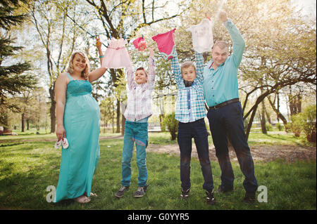 Happy pregnant couple with two sons holding girls' baby clothes on string  background spring nature Stock Photo