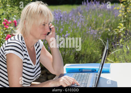 Side view of blond middle aged woman calling by smart phone and writing in a open notebook on the table. Stock Photo