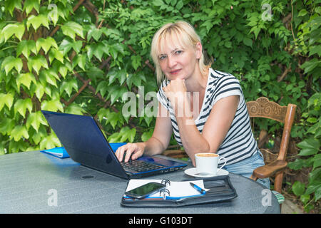 Pensive blond middle aged woman is sitting outdoors on the terrace  with open notebook, diary and smart phone on the table. Stock Photo