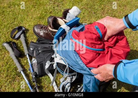 A hiker packs a red waterproof jacket into a blue hiking rucksack preparing to go for a hike. England, UK, Britain Stock Photo