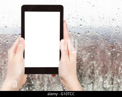 season concept - man photographs forest through window with rain drops in overcast autumn day on tablet pc with cut out screen w
