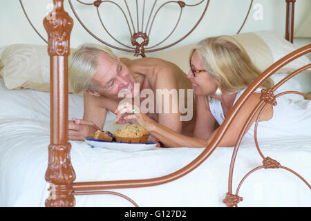 Retired man and woman lying in bed eating muffins for breakfast. Stock Photo
