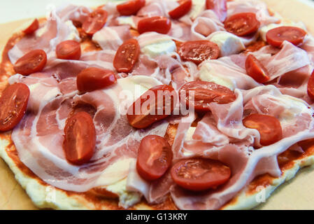 Closeup of a home made raw pizza with cheese, pancetta, cherry tomatoes and sauce on a wooden peel Stock Photo