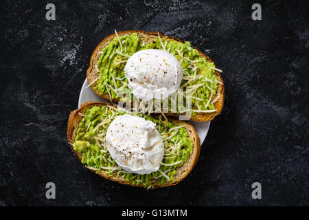 Two toasts with mashed avocado, cheese and poached egg on dark textured backdrop, top view. Healthy breakfast or snack Stock Photo
