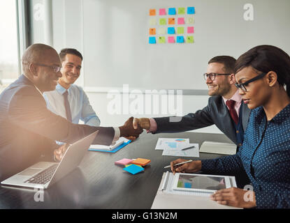 Multiracial team concluding a business agreement reaching across the table to shake hands with pleased smiles with focus to a yo Stock Photo