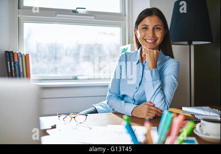 Cheerful woman with hand on chin wearing blue shirt while seated at desk covered with papers, coffee and eyeglasses and window i Stock Photo