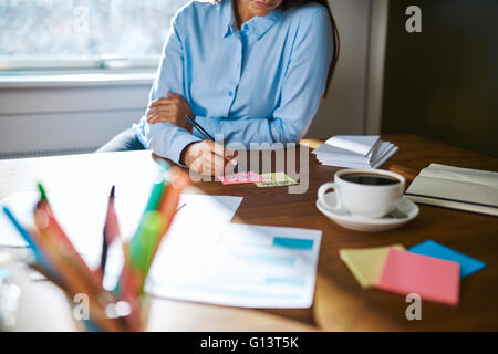 Female small business owner working from home sitting at her desk making notes on sticky memo cards , close up view with focus t