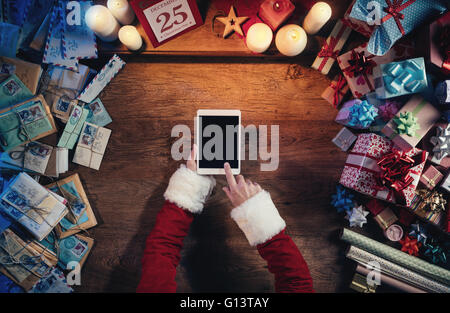 Santa Claus sitting at his desk and using a digital touch screen tablet surrounded by colorful Christmas gifts Stock Photo