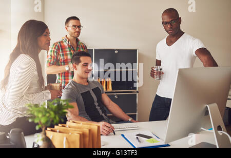 Diverse group of multi-ethnic young adult business people assembled around desk looking at something on their computer monitor Stock Photo