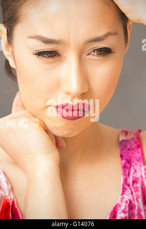 Agitated young woman head and shoulders portrait on gray background, with hand on forehead, looking puzzled, in red top. Stock Photo