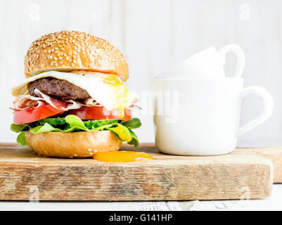 Breakfast set. Homemade beef burger with fried egg and vegetables and coffee cups on wooden board, white painted background. Sel Stock Photo