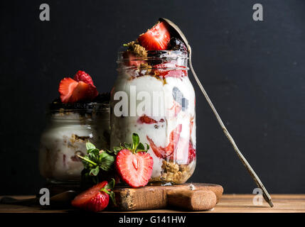 Yogurt oat granola with strawberries, mulberries, honey and mint leaves in tall glass jar on black backdrop, selective focus, co Stock Photo