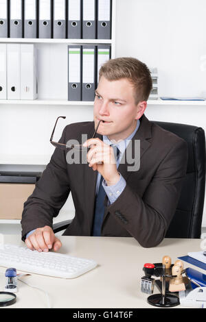 Young businessman with glasses is thoughtfully sitting in front of a computer keyboard at the desk in the office. A shelf is in Stock Photo