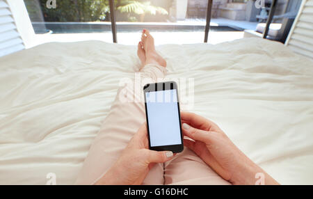 Closeup image of woman lying on a bed holding a smart phone with blank screen. POV shot of woman relaxing in bedroom using touch Stock Photo