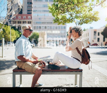 Side view of mature man sitting on bench and woman taking his photos with digital camera. Senior couple having fun on their vaca Stock Photo