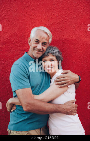 Portrait of beautiful senior couple embracing against red background. Loving mature couple standing together. Stock Photo