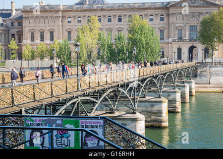 Pedestrians cross the River Seine in Paris on the Pont des Arts, it's railings covered in padlocks. Louvre museum in background. Stock Photo
