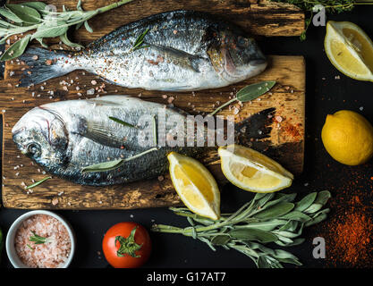 Fresh uncooked dorado or sea bream fish with lemon, herbs, oil, vegetables and spices on rustic wooden board over black backdrop Stock Photo