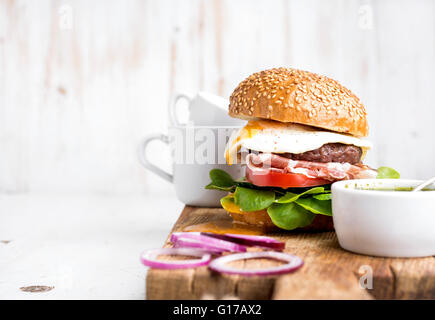 Breakfast set. Homemade beef burger with fried egg and vegetables, onion rings and coffee cups on wooden board, white painted ba Stock Photo