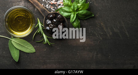 Greek black olives, fresh green sage, rosemary, basil herbs, salt and oil on dark rustic wooden background.  Top view, copy spac Stock Photo