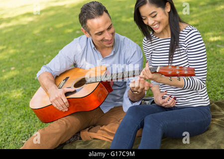 Couple sitting on grass playing acoustic guitar smiling Stock Photo