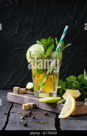 Glass of Iced green tea with lime, lemon, mint and sugar cubes on wooden chopping board over old wooden table. Dark rustic style Stock Photo