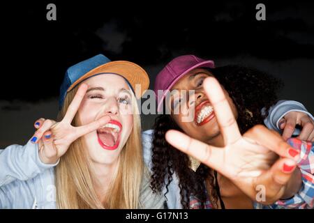 Young woman wearing baseball cap arm around friend looking at camera doing peace sign Stock Photo