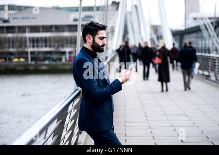 Businessman leaning against footbridge railing reading smartphone text, London, UK Stock Photo