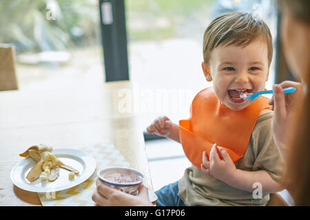 Happy baby boy sitting at table being fed yogurt by mother Stock Photo