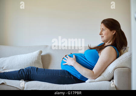 Side view of pregnant woman holding stomach, relaxing on sofa looking away smiling Stock Photo