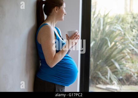 Side view of pregnant woman leaning against wall holding coffee cup looking away out of window smiling Stock Photo