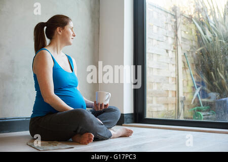 Pregnant woman sitting on floor crossed legged holding coffee cup looking away out of window Stock Photo