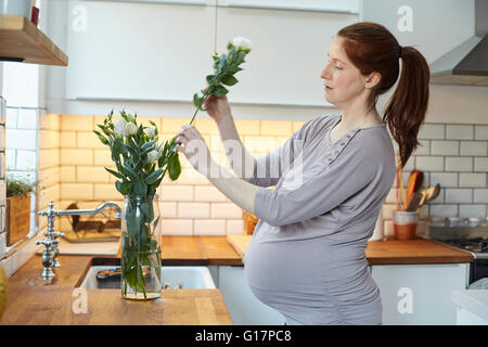 Side view of pregnant woman in kitchen arranging flowers in vase Stock Photo