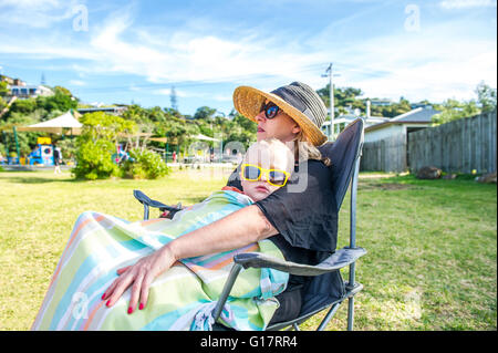 Boy wearing yellow sunglasses sitting on mother lap in park, Waiheke Island, New Zealand Stock Photo