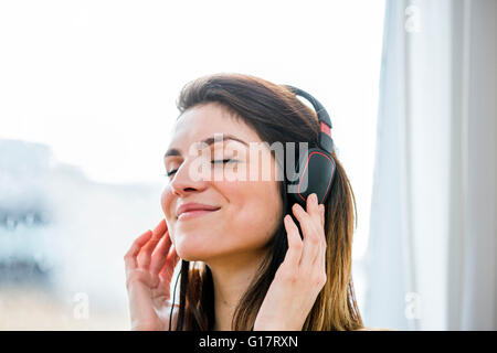Beautiful young woman listening to headphone music in front of  apartment window Stock Photo
