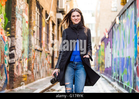 Portrait of stylish young woman with DSLR strolling along graffiti alley Stock Photo