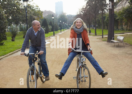 Couple riding bicycles on tree lined path in park, smiling Stock Photo