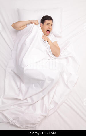 Vertical top view shot of a scared young man laying in bed covered with blanket and screaming Stock Photo