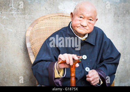 Old bald Chinese man in a blue coat sitting with a walking stick cane in the shadow on a chair outdoors on the street Stock Photo