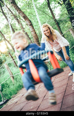 Mother and child outdoors in playground riding a  swing and smiling Stock Photo